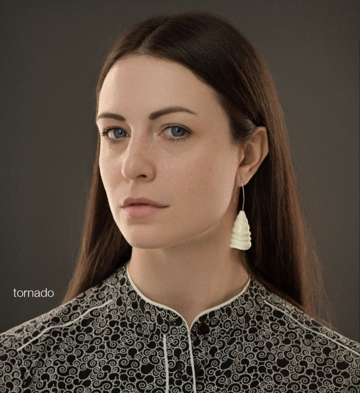 A woman with blue eyes and long dark hair wears the ‘Tornado’ porcelain earring by Serhiy Verbicki, paired with a black and white patterned blouse.