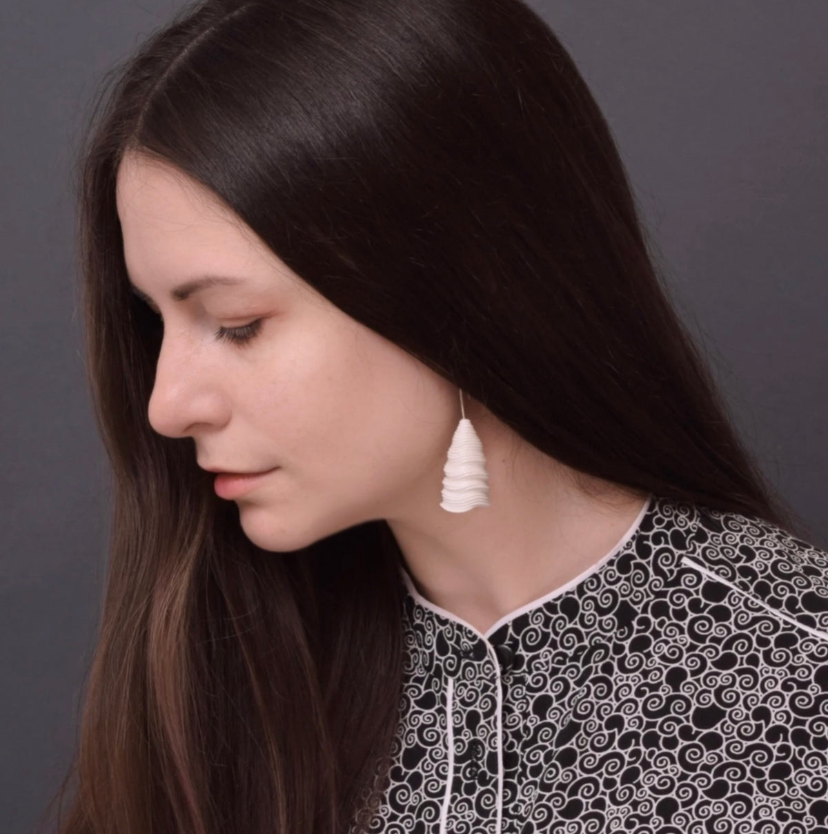 A woman with long dark hair wears the ‘Tornado’ porcelain earring by Serhiy Verbicki, paired with a patterned black and white blouse.