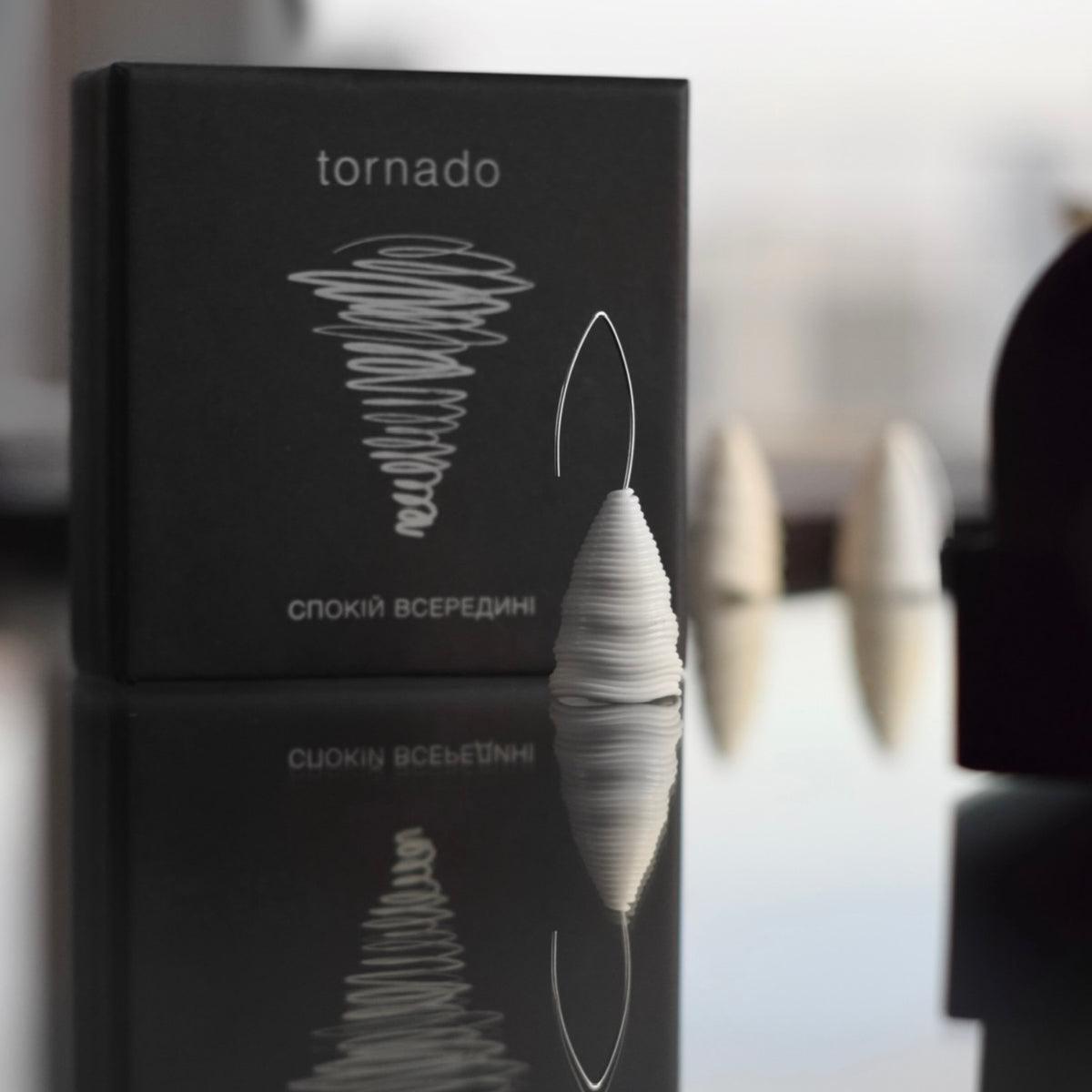 The ‘Tornado’ porcelain earring by Serhiy Verbicki, standing beside its black packaging with the slogan 'Calm Inside,' reflected on a glossy surface.